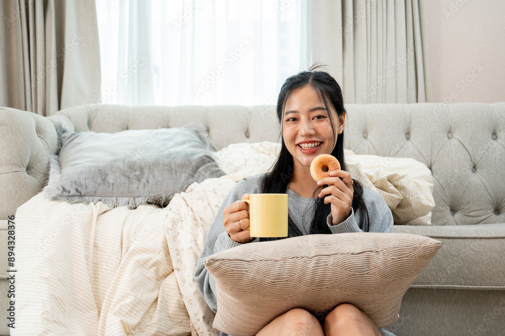 A charming Asian woman is having breakfast in the living room, eating a donut and drinking coffee. A charming Asian woman is having breakfast in the living room, eating a donut and drinking coffee.