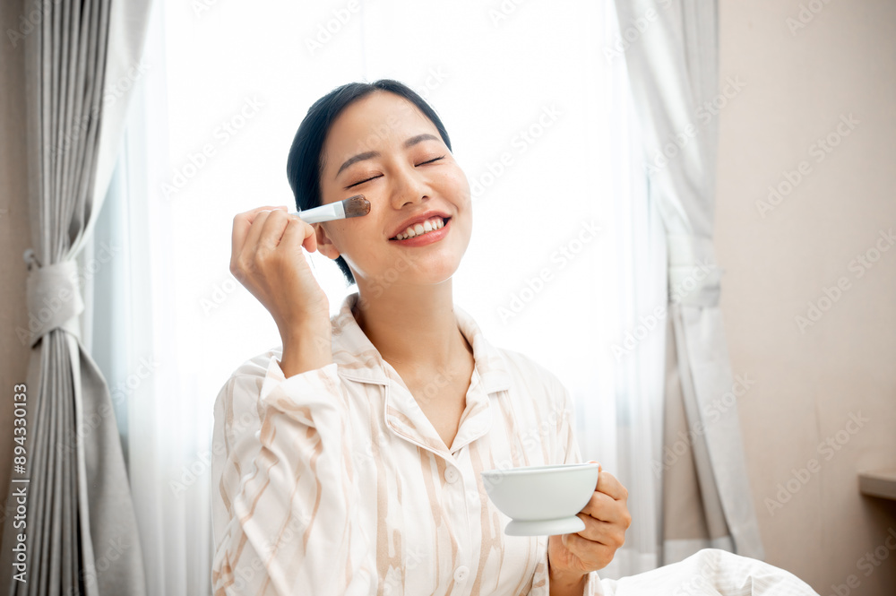 A beautiful, smiling young Asian woman in pajamas is applying a facial treatment mask to her face. A beautiful, smiling young Asian woman in pajamas is applying a facial treatment mask to her face.