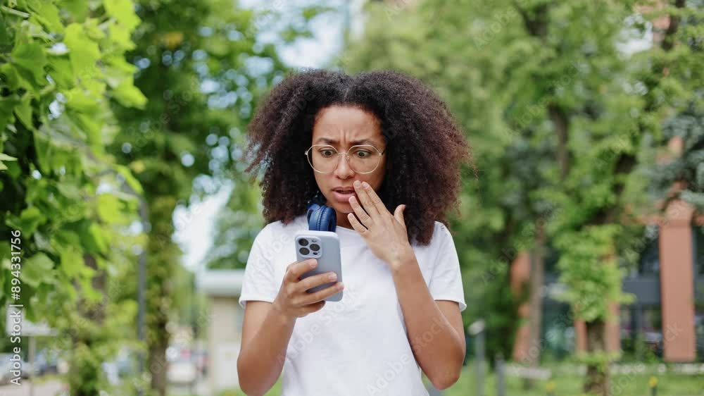 Startled african american woman with wireless headphones around neck looking on personal smartphone and feeling surprised. Amazed young woman gestures with hands and experiencing fear outdoors. Startled african american woman with wireless headphones around neck looking on personal smartphone and feeling surprised. Amazed young woman gestures with hands and experiencing fear outdoors.