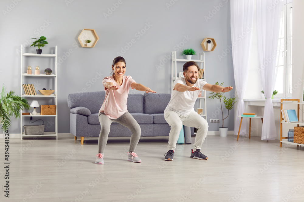 Obraz premium Happy sporty young couple wearing sportswear having sport workout in living room at home. Cheerful man and woman doing squats exercises together. Concept of family sport and fitness.