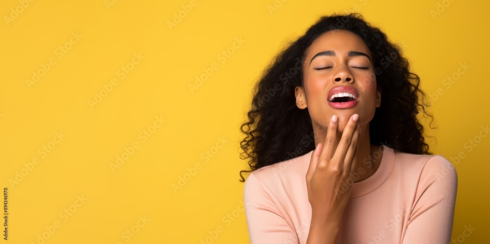 25-year-old female, African-American, yawning with her hand covering her mouth, against a pastel yellow background 25-year-old female, African-American, yawning with her hand covering her mouth, against a pastel yellow background