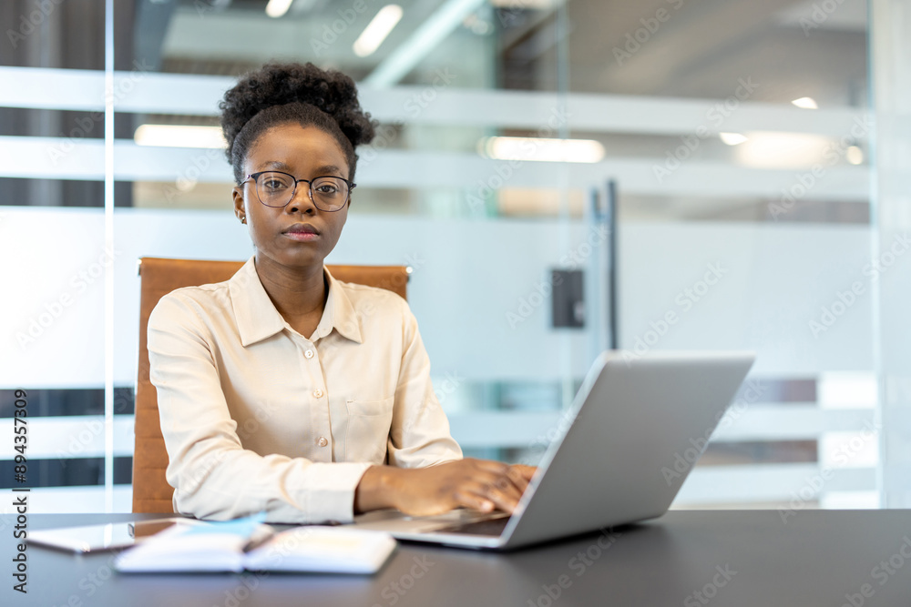Confident businesswoman with glasses sitting at desk, working on laptop in modern office. Professional attire, focused expression, depicts dedication and productivity in corporate environment Confident businesswoman with glasses sitting at desk, working on laptop in modern office. Professional attire, focused expression, depicts dedication and productivity in corporate environment