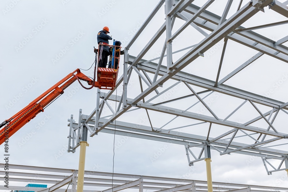 Fototapeta premium Man worker on a crane performs high-rise work on welding metal structures of new tower at height
