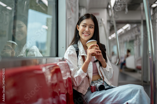 A happy young Asian female tourist is sipping coffee while sitting on a seat in a sky train.