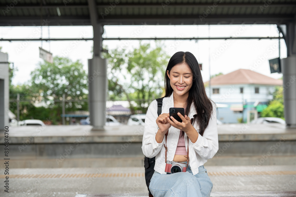 Positive Asian female traveler is using her smartphone while sitting on a bench at a train platform. Positive Asian female traveler is using her smartphone while sitting on a bench at a train platform.