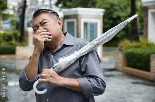 Asian middle-aged man carrying umbrella after raining, ill and sick Asian uncle having nausea, stomach upset, allergic stomach, vomit symptom, queasiness, emesis photo