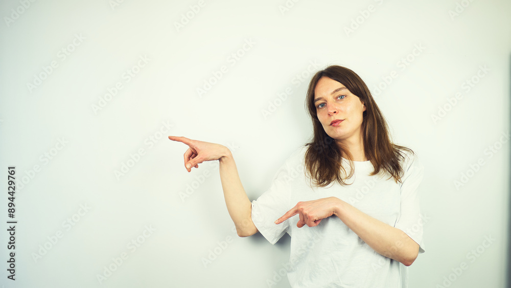 Portrait of beautiful Caucasian young female model with blue eyes and white shirt pointing her finger, direction gesture, isolated on white background, copy space area