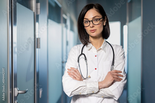 Confident female doctor with glasses and stethoscope standing in hospital corridor. Professional medical staff in healthcare environment. Concept of medical expertise, healthcare, and hospital work. photo