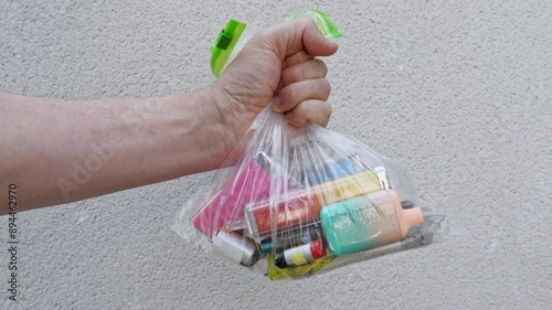 A white male grips a clear plastic bag with a collection of discarded vapes inside. The bag is held in front of a pale coloured stone wall.