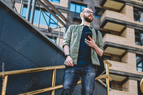 Pondering bearded young man looking away while standing on stairs with smartphone in hands in urban setting.Thoughtful hipster guy dressed in casual wear thinking on route holding cellular device