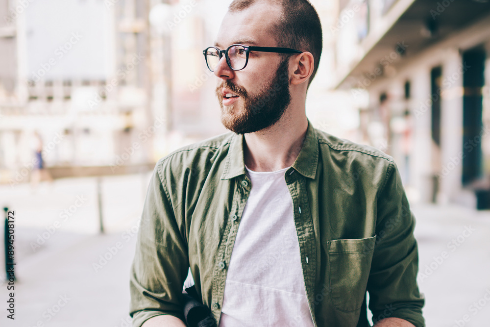 Caucasian young man in eyeglasses looking away while sitting outdoors and holding modern smartphone device in hands.Pondering hipster guy installing app on mobile phone using 4G internet connection Caucasian young man in eyeglasses looking away while sitting outdoors and holding modern smartphone device in hands.Pondering hipster guy installing app on mobile phone using 4G internet connection