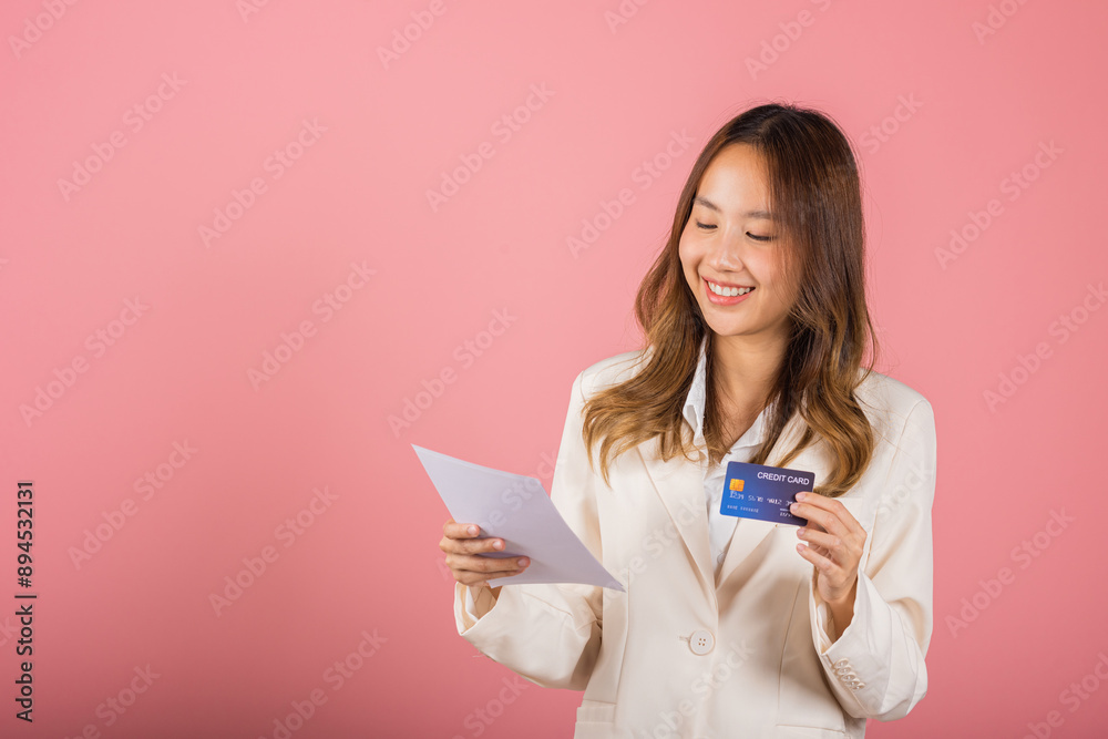 Fototapeta premium Portrait of happy Asian young woman holding paper calculating bills on hand and credit card for payment, female person she hold bill or paperwork, studio shot isolated on pink background