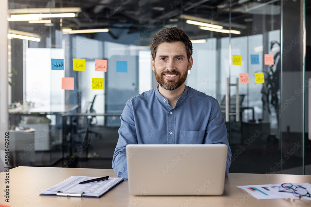 Portrait of a smiling young man working in a modern office using a laptop, sitting at a desk and looking at the camera Portrait of a smiling young man working in a modern office using a laptop, sitting at a desk and looking at the camera