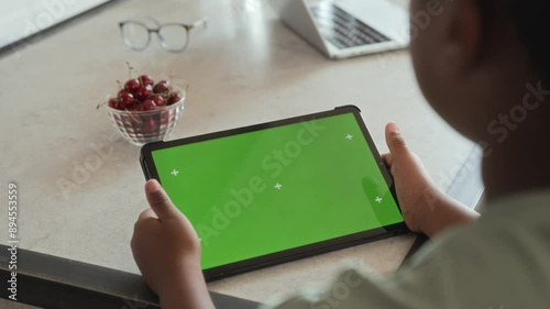 African American boy is holding in hands and using tablet pad with green screen chroma key display and motion trackers, sitting at table