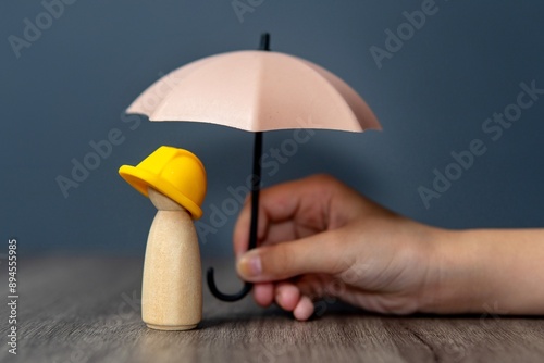 Closeup image of hand holding a umbrella over a wooden doll wearing a yellow hard hat. Worker and employee insurance concept.
