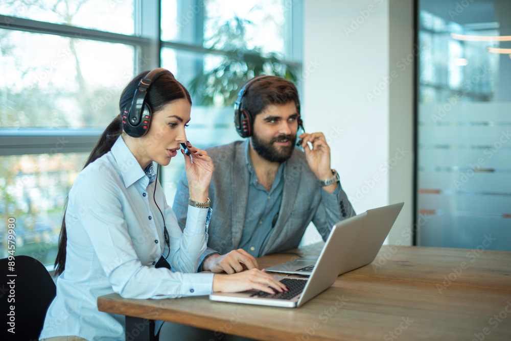 Female and male operators in headsets using laptops while working together in office. Female and male operators in headsets using laptops while working together in office.