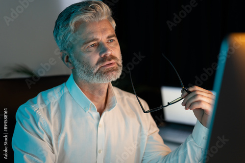 Фотография Thoughtful Senior Man Working Late Night In Office, Holding Glasses And Looking