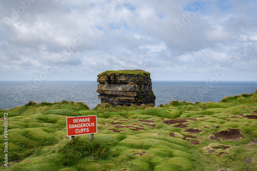 a red beware sign on the edge of a grassy cliff in Ireland with a sea stack rock formation
