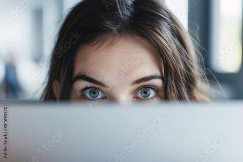 A woman peeks over the top of a laptop screen with only her curious blue eyes and forehead visible photo