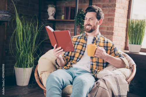 Photo of handsome good mood guy wear plaid shirt smiling having rest reading story drink tea indoors room home house