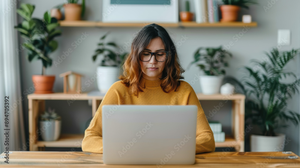 Woman wearing glasses working on a laptop at a wooden desk with plants and bookshelves in the background, creating a cozy workspace vibe.
