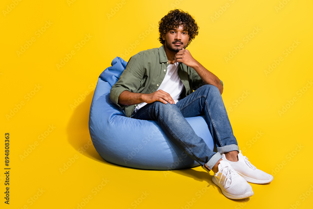 Portrait photo of young funny mexican guy in khaki shirt sitting pouf at lounge seriously touch chin isolated on yellow color background Portrait photo of young funny mexican guy in khaki shirt sitting pouf at lounge seriously touch chin isolated on yellow color background