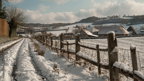 A picturesque rural scene with a snow-covered path, wooden fence, and fields, leading to a village under a cloudy sky