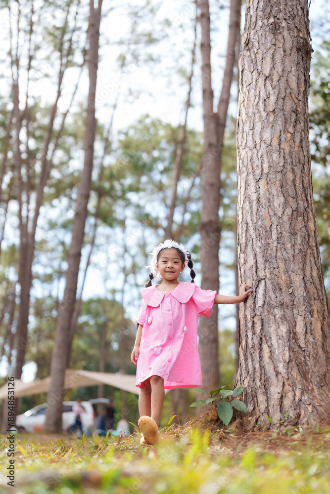 Asian girl in pink dress standing near tree in park, smiling and enjoying nature. Concept of childhood happiness, innocence, and playful exploration in a serene, sunlit forest setting. Asian girl in pink dress standing near tree in park, smiling and enjoying nature. Concept of childhood happiness, innocence, and playful exploration in a serene, sunlit forest setting.
