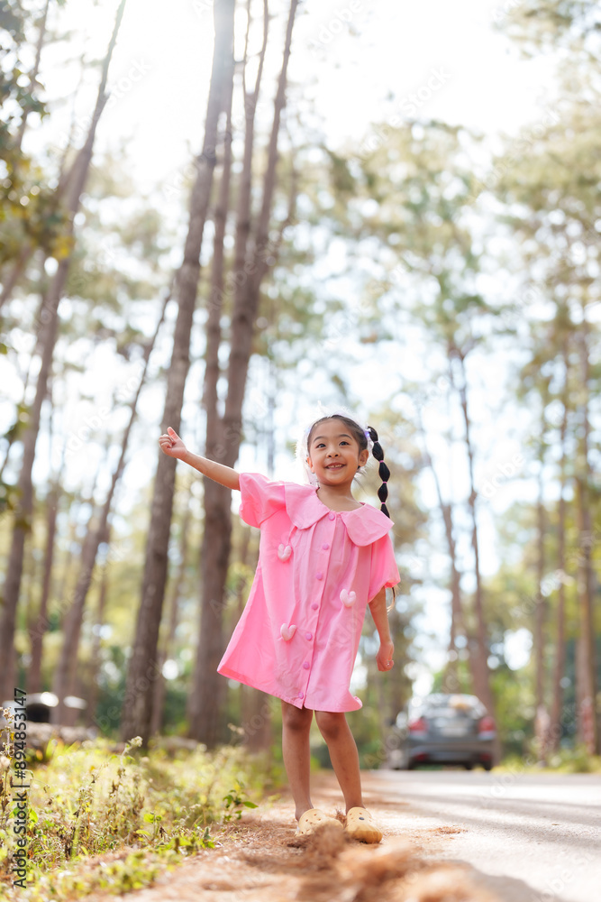 Asian girl in pink dress pointing excitedly in park with trees and car in background. Concept of childhood joy, discovery, and carefree moments in a sunlit natural setting. Asian girl in pink dress pointing excitedly in park with trees and car in background. Concept of childhood joy, discovery, and carefree moments in a sunlit natural setting.