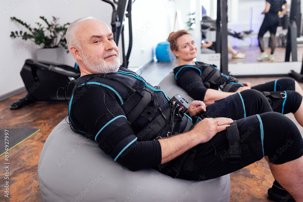 Obraz premium Elderly couple sits on massage chairs while waiting for ems workout in gym or fitness club.