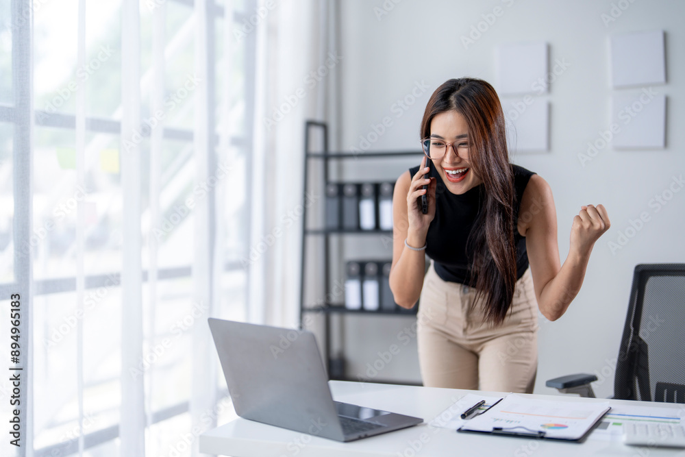 A woman is standing at a desk with a laptop and a cell phone A woman is standing at a desk with a laptop and a cell phone