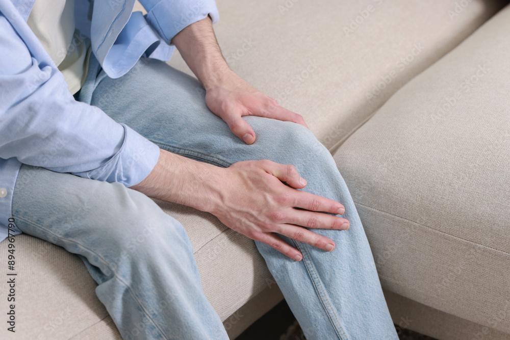 Man suffering from knee pain on sofa indoors, closeup Man suffering from knee pain on sofa indoors, closeup