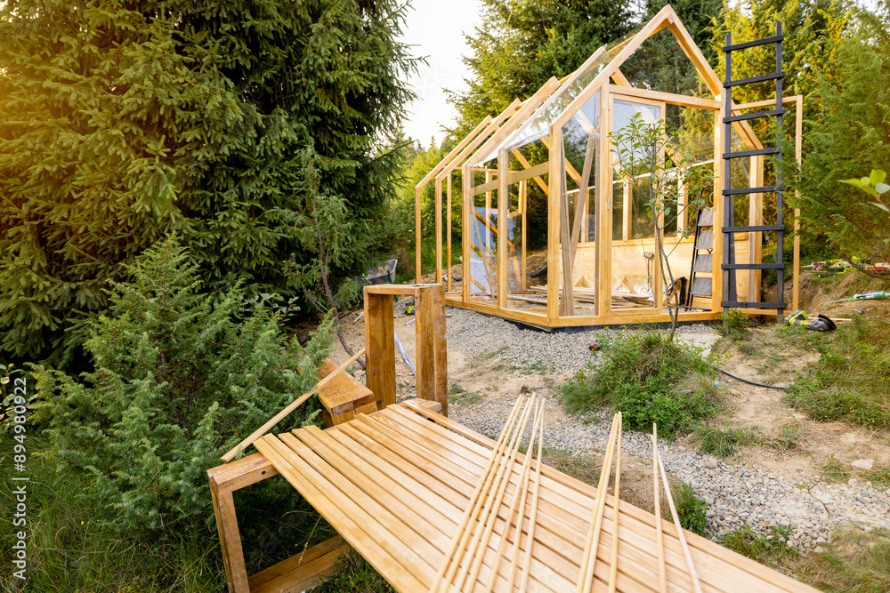 A wooden greenhouse under construction in a lush backyard surrounded by tall trees. The scene captures the early stages of a DIY project in an eco-friendly environment, highlighting sustainable living A wooden greenhouse under construction in a lush backyard surrounded by tall trees. The scene captures the early stages of a DIY project in an eco-friendly environment, highlighting sustainable living