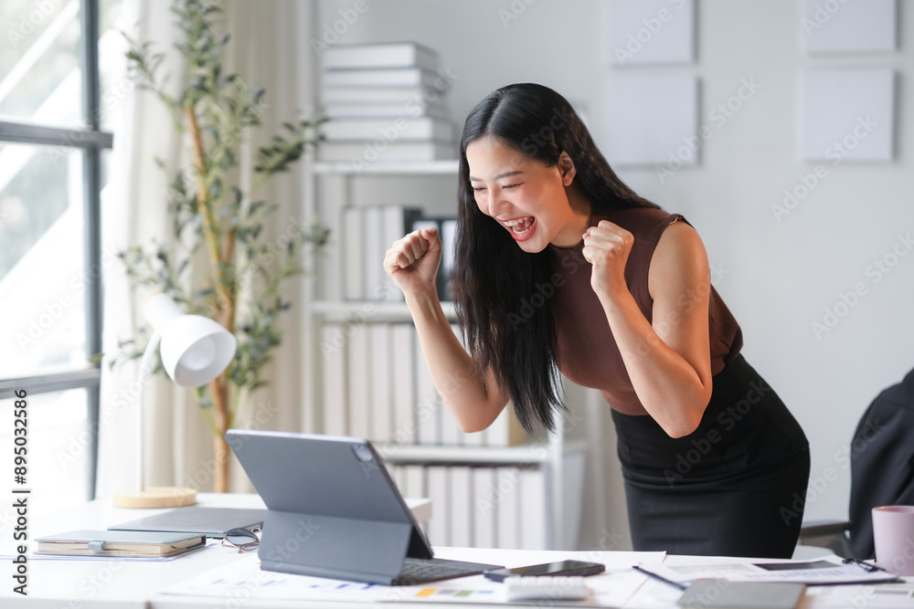 Young asian businesswoman is working in a modern office. She is very happy and excited from looking at her laptop, raising her hands with joy Young asian businesswoman is working in a modern office. She is very happy and excited from looking at her laptop, raising her hands with joy