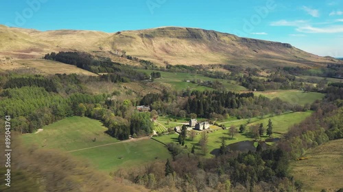 Aerial View Flying Over Pine Tree Forest to Reveal Countryside Castle in a Scottish Valley, with Lush Green Fields, Pine Forest and Rolling Hills Behind in the British Countryside photo