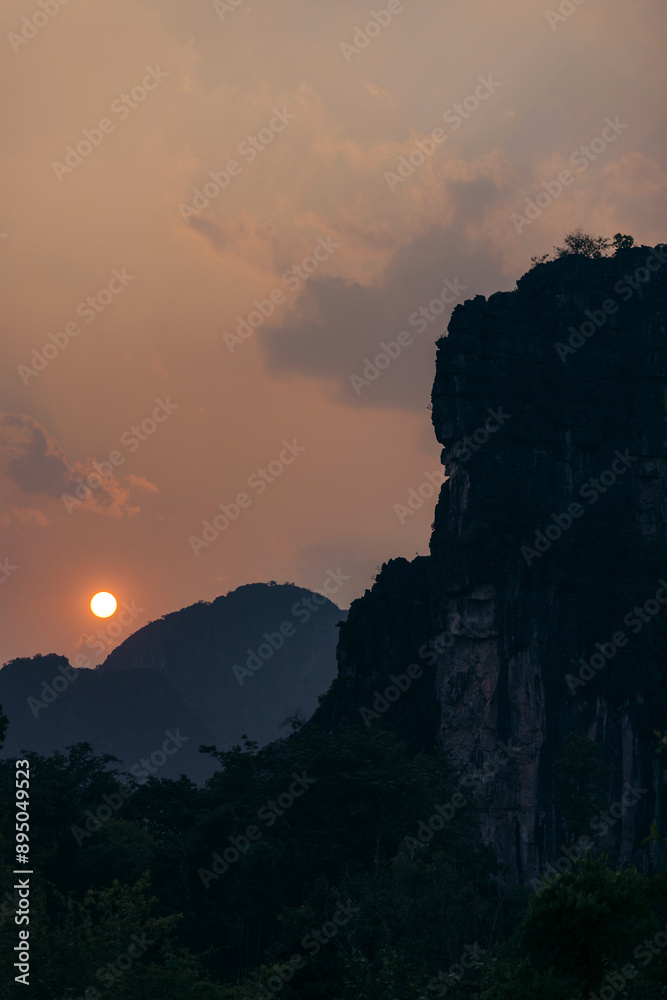 Sun setting in a warmly lit sky behind silhouetted karst formations in the Thakhek area of Laos; Khammouane Province, Laos Sun setting in a warmly lit sky behind silhouetted karst formations in the Thakhek area of Laos; Khammouane Province, Laos