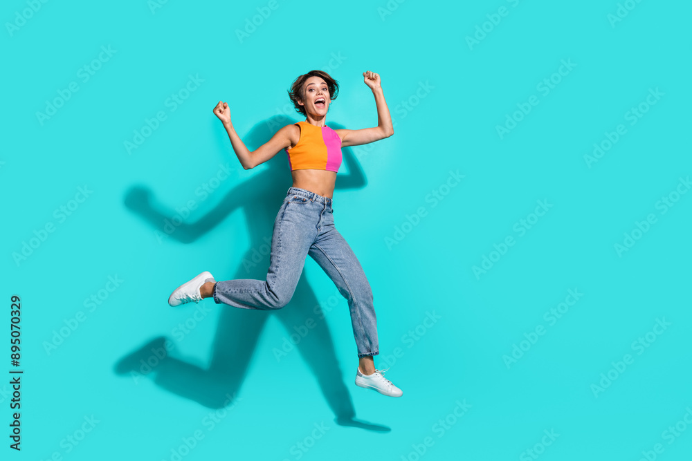 Full length photo of pretty lucky woman wear pink orange top jumping high rising fists empty space isolated teal color background Full length photo of pretty lucky woman wear pink orange top jumping high rising fists empty space isolated teal color background