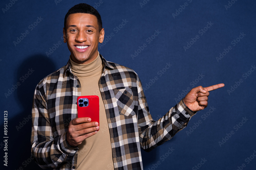 Photo portrait of handsome young man point empty space hold device dressed stylish plaid garment isolated on dark blue color background Photo portrait of handsome young man point empty space hold device dressed stylish plaid garment isolated on dark blue color background