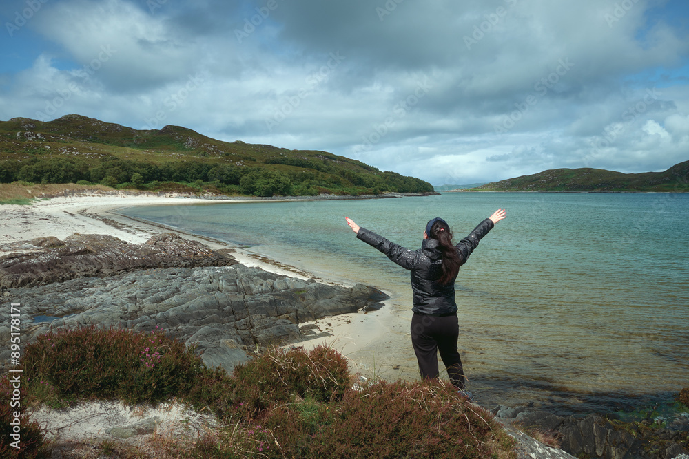 A woman from the back on the shore of the sea bay raised her hands in the air rejoicing in the beautiful scenery. West Coast, Scotland A woman from the back on the shore of the sea bay raised her hands in the air rejoicing in the beautiful scenery. West Coast, Scotland