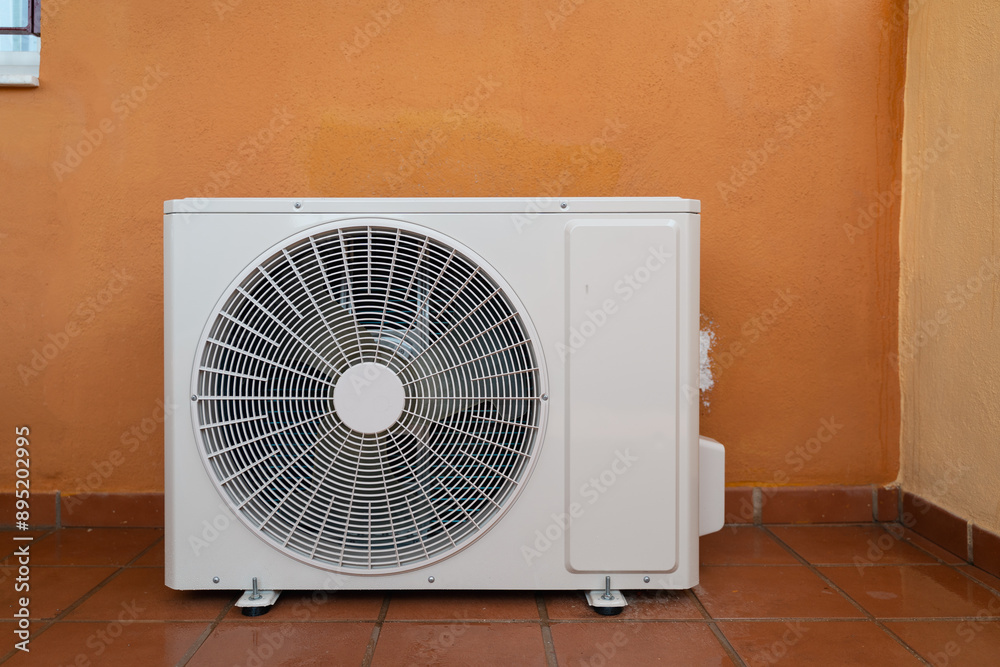A white air conditioner unit is mounted on a balcony wall against an orange background. Extreme heat, temperature control at home concept A white air conditioner unit is mounted on a balcony wall against an orange background. Extreme heat, temperature control at home concept