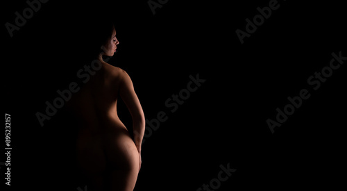 A nude woman is standing in the darkness, illuminated by a faint light source, casting shadows around her. Her figure is silhouetted against the black background.
