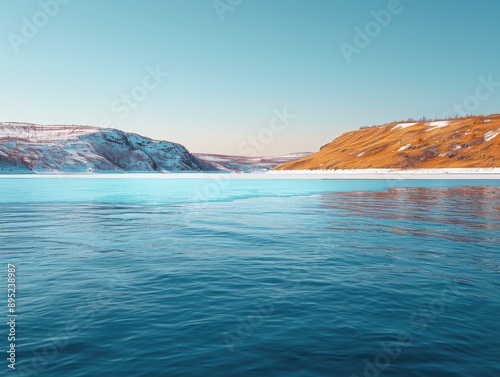 A serene view of a frozen lake with clear blue water in Lapland, Murmansk Oblast, Russia, surrounded by snow-capped mountains and brown hills under a clear sky.