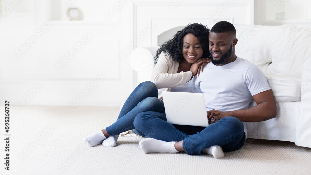 A couple smiles at a laptop while sitting on the floor in their home. A couple smiles at a laptop while sitting on the floor in their home.