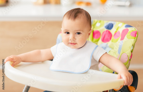 A cute baby wearing a white bib sits comfortably in a high chair in a well-lit kitchen. The baby looks curious and content while holding onto the edge of the high chair.