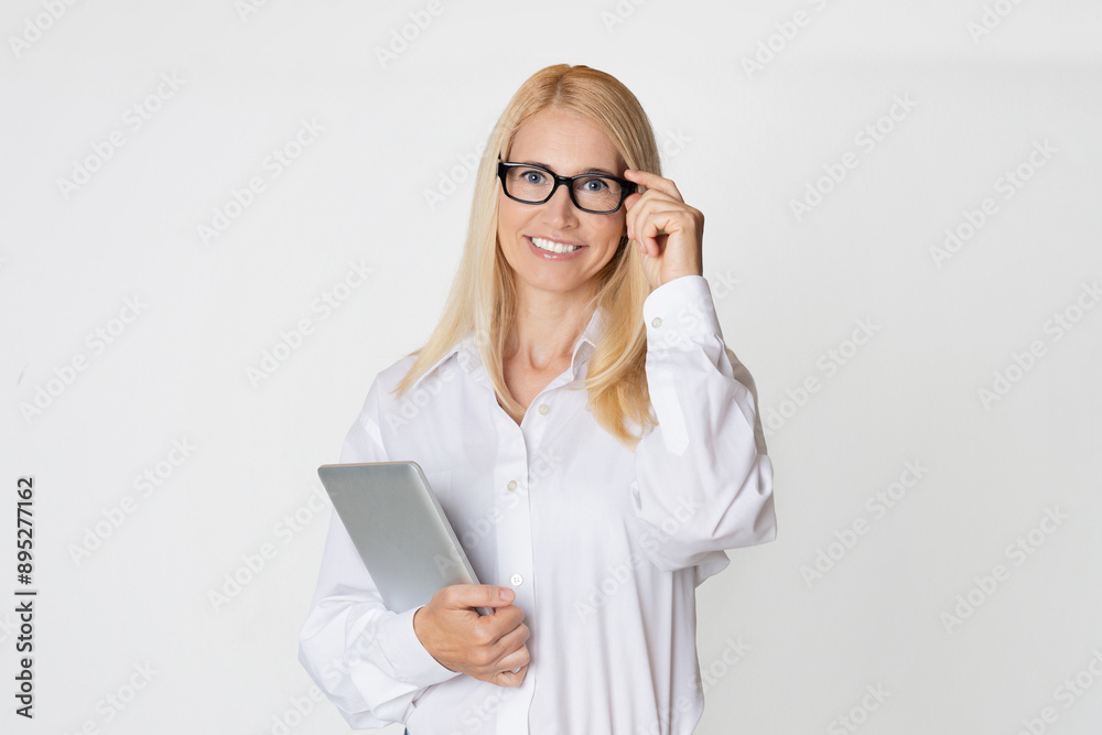 A young, attractive businesswoman wearing glasses smiles and looks directly at the camera. She is holding a tablet computer in her left hand and adjusting her glasses with her right hand. A young, attractive businesswoman wearing glasses smiles and looks directly at the camera. She is holding a tablet computer in her left hand and adjusting her glasses with her right hand.