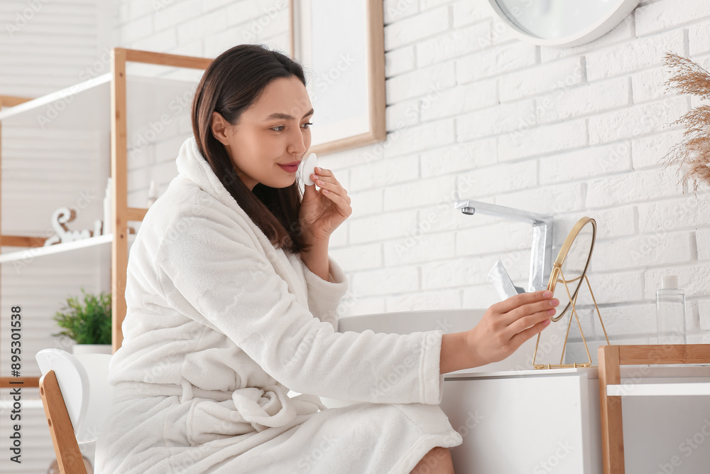 Beautiful young woman removing makeup with cotton pad near mirror in bathroom Beautiful young woman removing makeup with cotton pad near mirror in bathroom