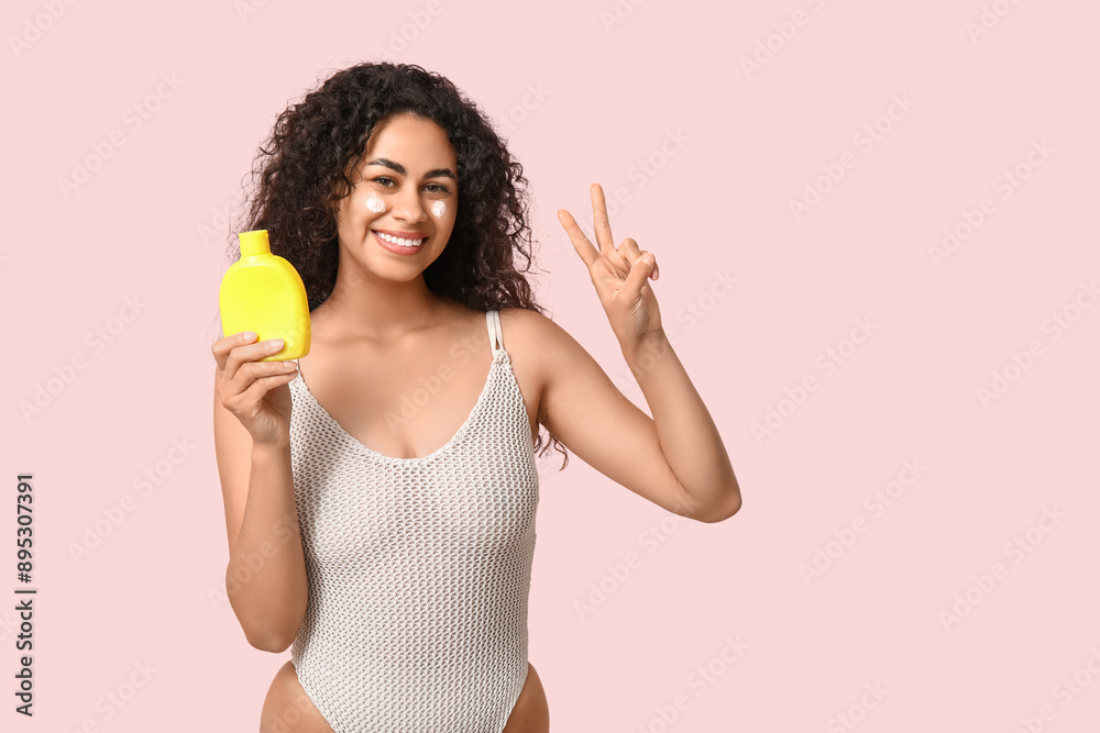 Beautiful young happy African-American woman with bottle of sunscreen cream showing victory gesture on pink background Beautiful young happy African-American woman with bottle of sunscreen cream showing victory gesture on pink background