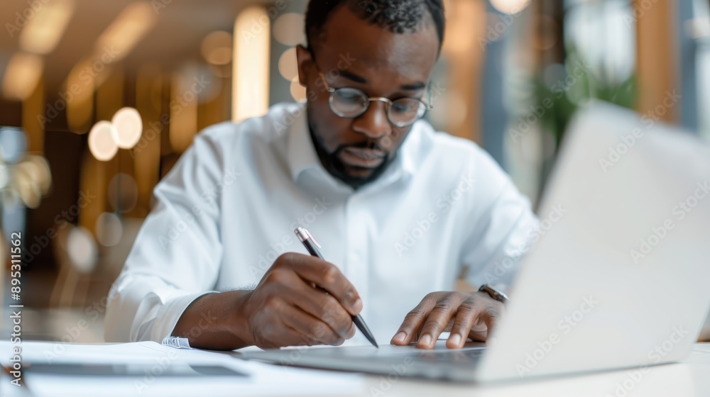 A diligent man works attentively on his laptop while taking notes with a pen, epitomizing focus and productivity in a modern workspace, surrounded by soft, ambient lighting. A diligent man works attentively on his laptop while taking notes with a pen, epitomizing focus and productivity in a modern workspace, surrounded by soft, ambient lighting.