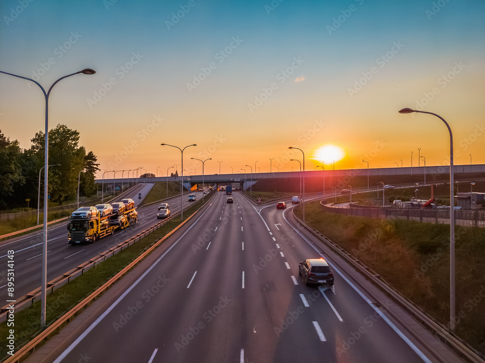 Obraz premium Passenger cars and a car transporter on a multi-line highway in the city at sunset.