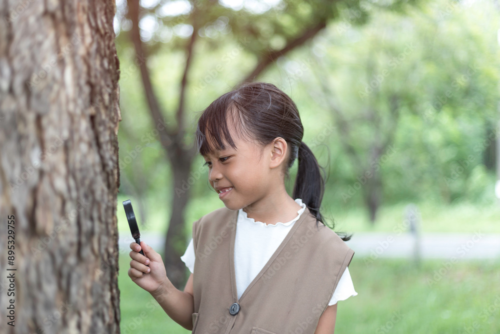 A girl, holding a magnifying glass, peers intently at the small creatures near the tree. A girl, holding a magnifying glass, peers intently at the small creatures near the tree.